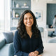 A professional portrait of a smiling South Asian woman in a bright, modern setting with soft blue and white decor accents.